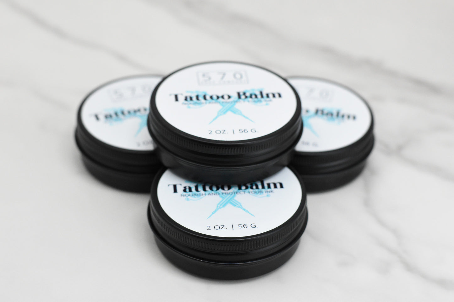 Five round containers of 570 Soap Co.'s Tattoo Balm - Nourish and Protect Your Ink, enriched with natural ingredients, are elegantly arranged on a white marble surface. Each 2 oz (56 g) container is designed to promote skin hydration, ensuring your tattoos remain vibrant and well-nourished.