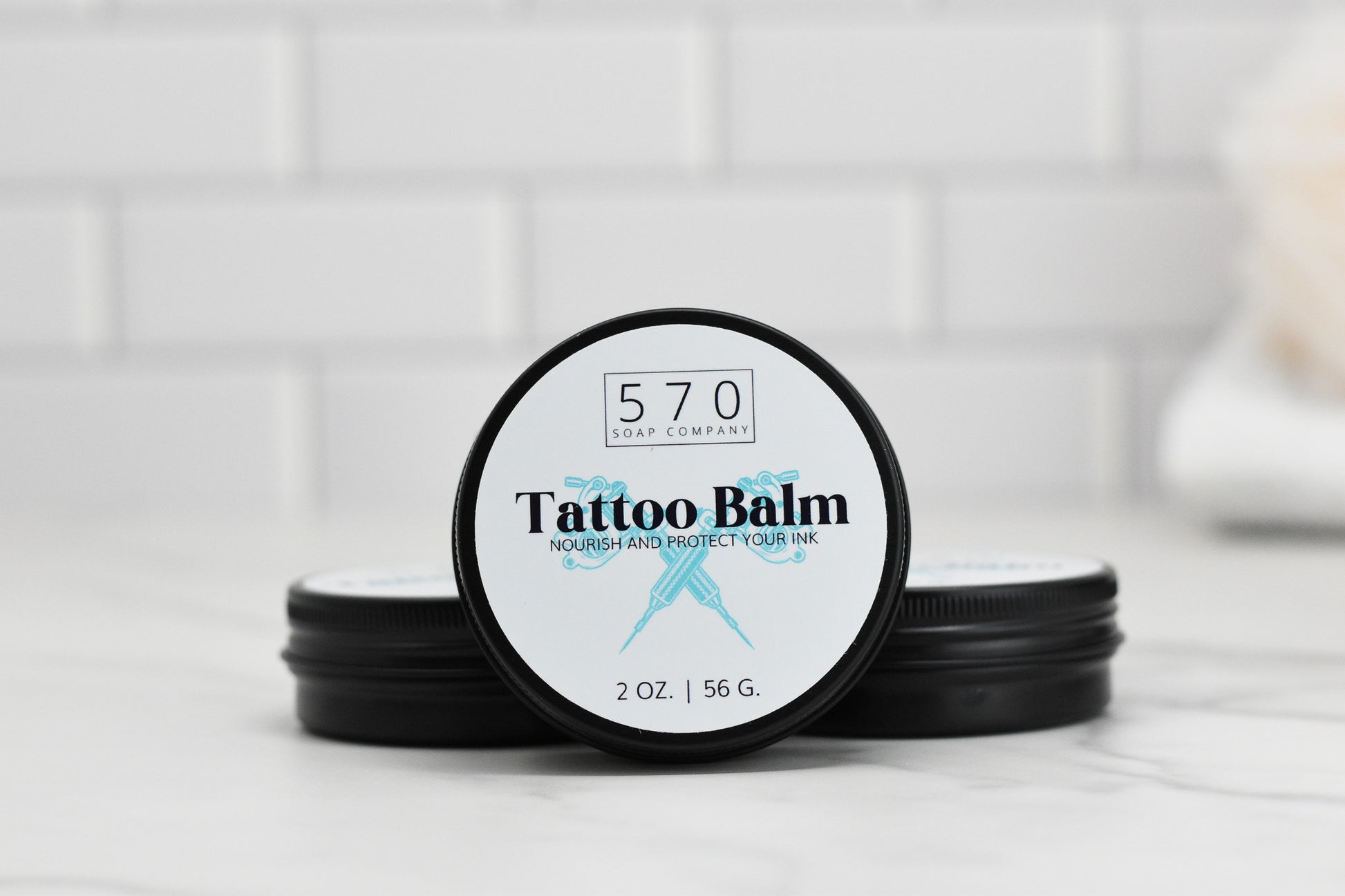 A tin of Tattoo Balm from 570 Soap Co. sits on a marble surface, providing skin hydration with its nourishing formula. Enclosed in a 2 oz (56 g) container, the label guarantees to nourish and protect your ink, as two additional tins appear in the background. Crafted with natural ingredients for optimal care.