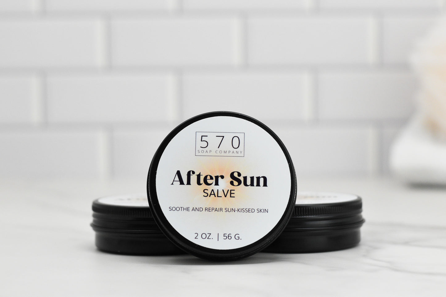 A small jar of 570 Soap Co.'s After Sun Salve is positioned against a minimalist white tile background. The label reads "After Sun Salve - Soothe and Repair Sun-Kissed Skin" with the contents listed as 2 oz / 56 g.