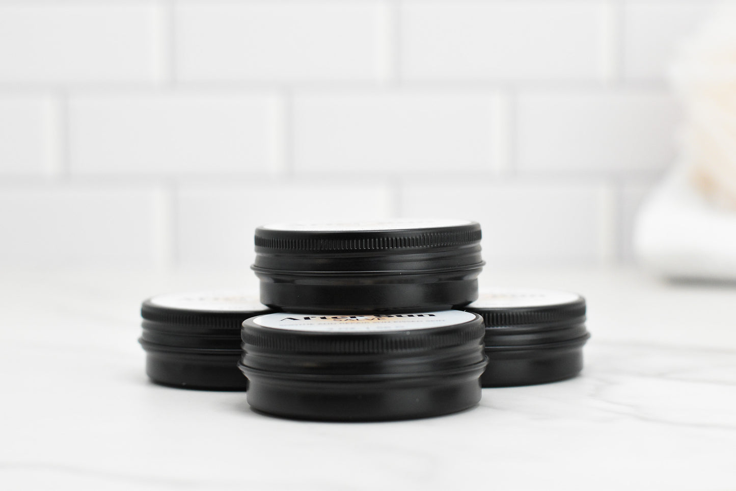 On a white marble surface, four compact black jars from 570 Soap Co., labeled "After Sun Salve - Soothe and Repair Sun-Kissed Skin," are stacked in pairs against a backdrop of white brick tiles. Designed specifically for sun-kissed skin, these jars promise soothing and reparative benefits, with their slightly raised lids inviting you to explore the magic inside.