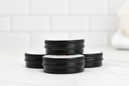 On a white marble surface, four compact black jars from 570 Soap Co., labeled "After Sun Salve - Soothe and Repair Sun-Kissed Skin," are stacked in pairs against a backdrop of white brick tiles. Designed specifically for sun-kissed skin, these jars promise soothing and reparative benefits, with their slightly raised lids inviting you to explore the magic inside.