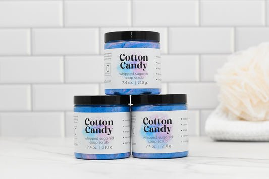 A trio of Cotton Candy Sugared Whipped Soap Scrub jars from 570 Soap Co. are stacked on a marble surface, their pastel tones contrasting beautifully against a white subway tile background. Perfect for exfoliation, the creamy formula sits next to a beige loofah on the right.