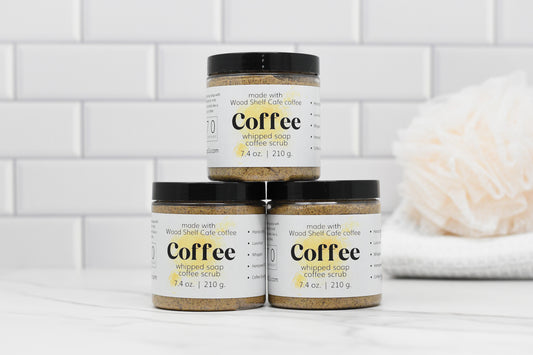 Three jars of 570 Soap Co.'s Whipped Soap Espresso Coffee Scrub are neatly stacked against a white tiled background. In the back to the right, an espresso grounds-infused bath sponge lies gently. The jars feature a simple, modern design with clear labeling and are enriched with nourishing apricot kernel oil.