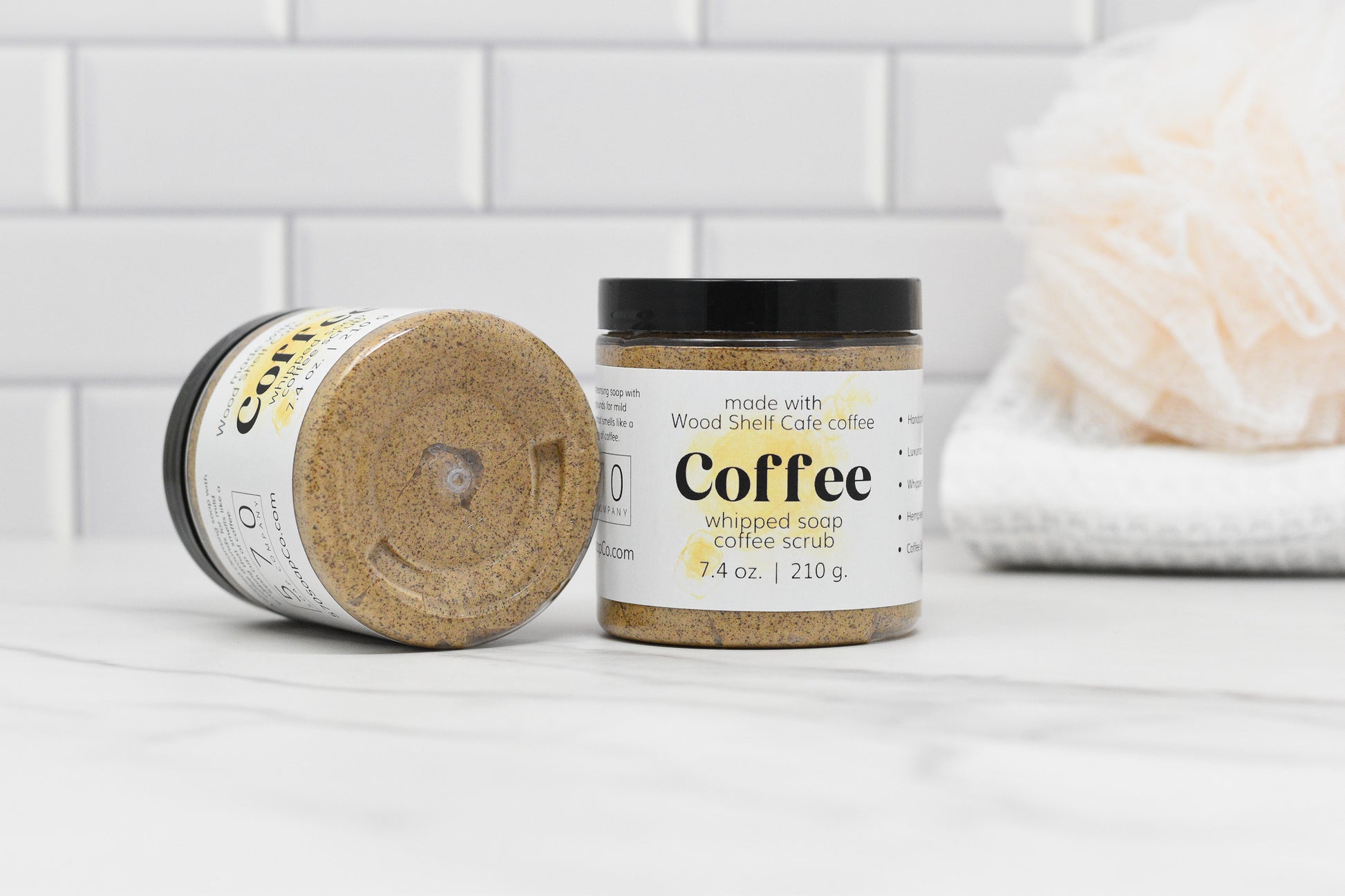 Two jars of Whipped Soap Espresso Coffee Scrub by 570 Soap Co., enhanced with apricot kernel oil, are positioned on a white marble surface against a tiled background. One jar is upright while the other is on its side. A white bath sponge and neatly folded towel contribute to the scene's elegance.