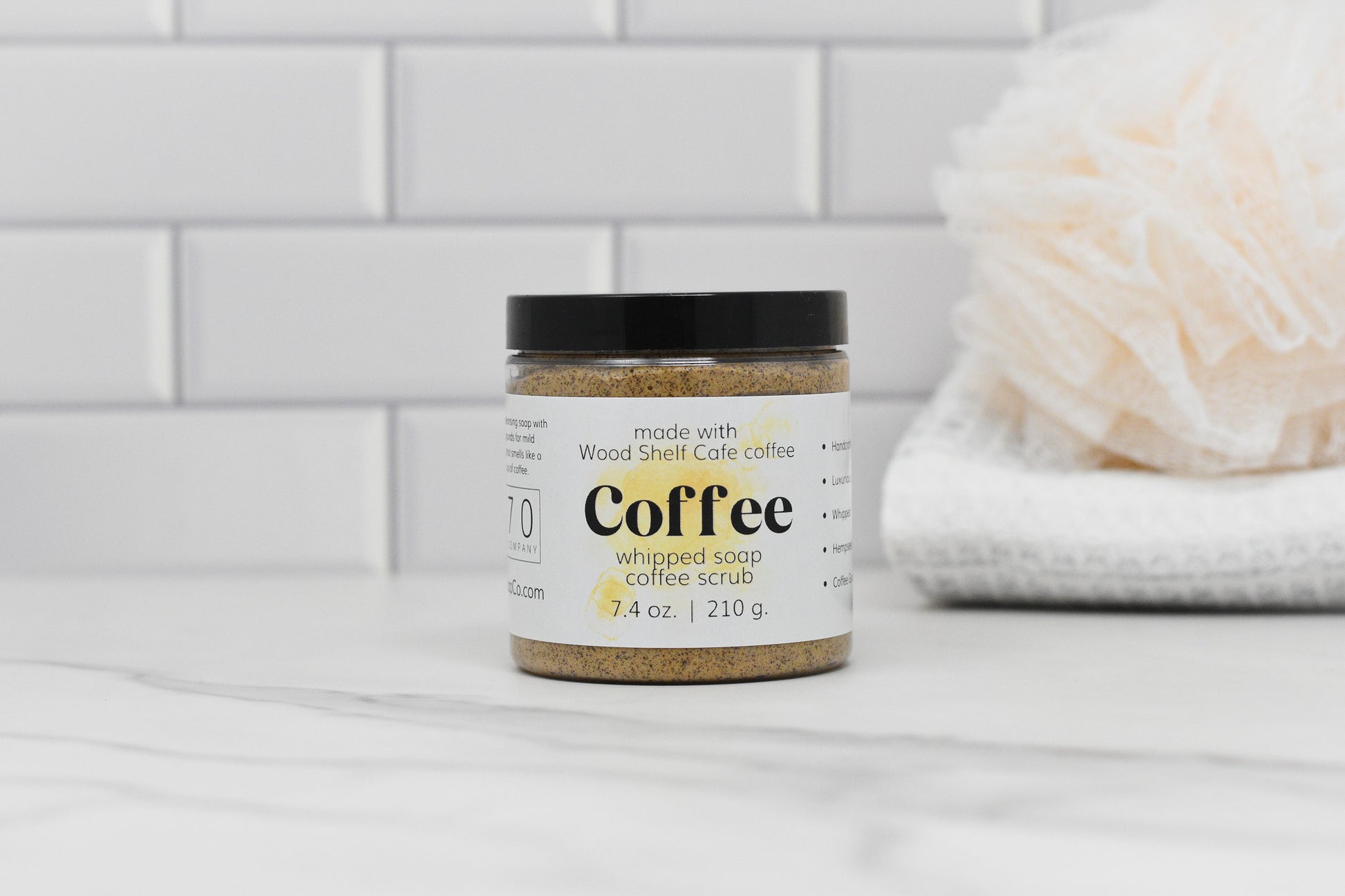 A jar of Whipped Soap Espresso Coffee Scrub, enriched with apricot kernel oil by 570 Soap Co., rests on a marble surface in front of a white tiled wall. The label highlights its inclusion of Wood Shelf Cafe coffee. In the background, a white bath pouf and a folded towel enhance the spa-like ambiance.