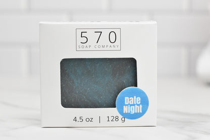 A bar of soap labeled Date Night Bar Soap: The Ultimate Man Soap, boasting an intricate blend of scents, peeks through the window of a white box. The packaging features 570 Soap Co. and indicates a weight of 4.5 oz or 128 g. The backdrop is light and blurred, inviting you on a fragrant journey.