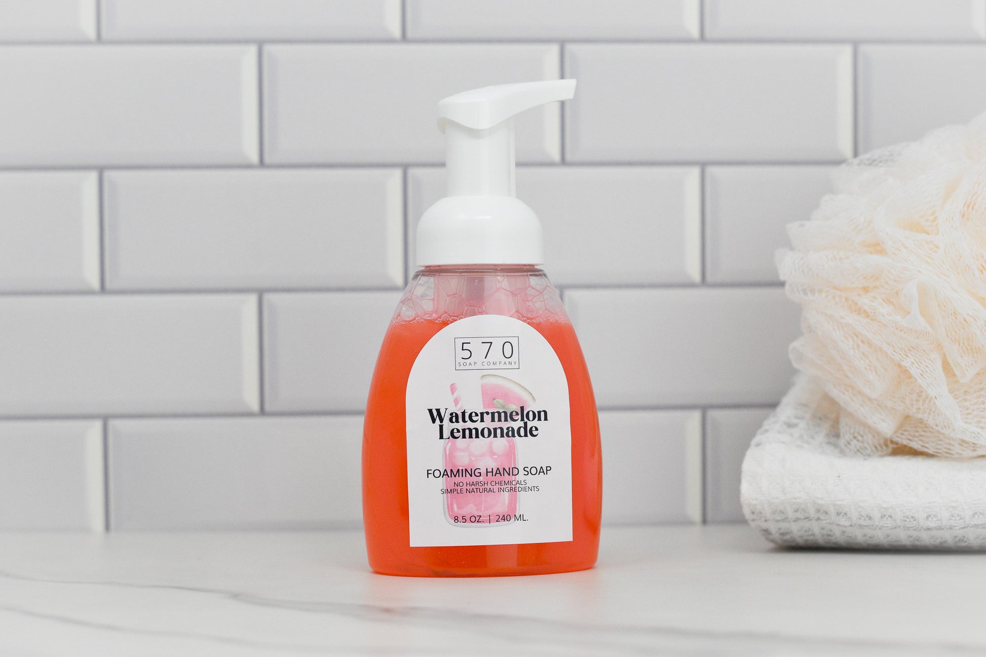 A bottle of 570 Soap Co.'s Watermelon Lemonade Scented Foaming Hand Soap is placed on a bathroom countertop, with a white tiled wall in the background. A loofah and folded towel are nearby, creating a clean, spa-like setting that highlights its moisturizing benefits.