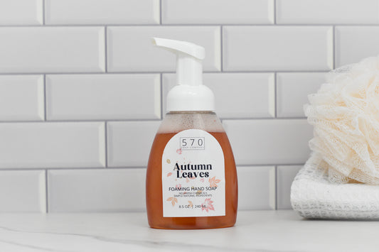 An Autumn Leaves Scented Foaming Hand Soap by 570 Soap Co., infused with a refreshing fall aroma, sits on a bathroom counter with a white tile backsplash. Next to it is a stack of folded white towels and a loofah.