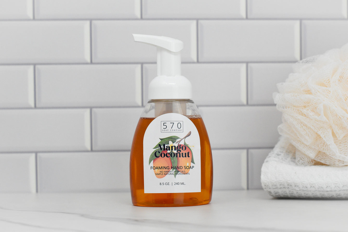 A bottle of 570 Soap Co.s Mango Coconut Scented Foaming Hand Soap, featuring a white pump and clear container, sits on a bathroom counter. In the background, a white folded towel and bath pouf rest against a white tiled wall.