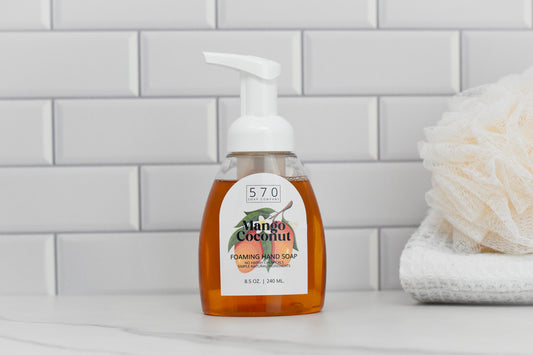 A bottle of 570 Soap Co.s Mango Coconut Scented Foaming Hand Soap, featuring a white pump and clear container, sits on a bathroom counter. In the background, a white folded towel and bath pouf rest against a white tiled wall.