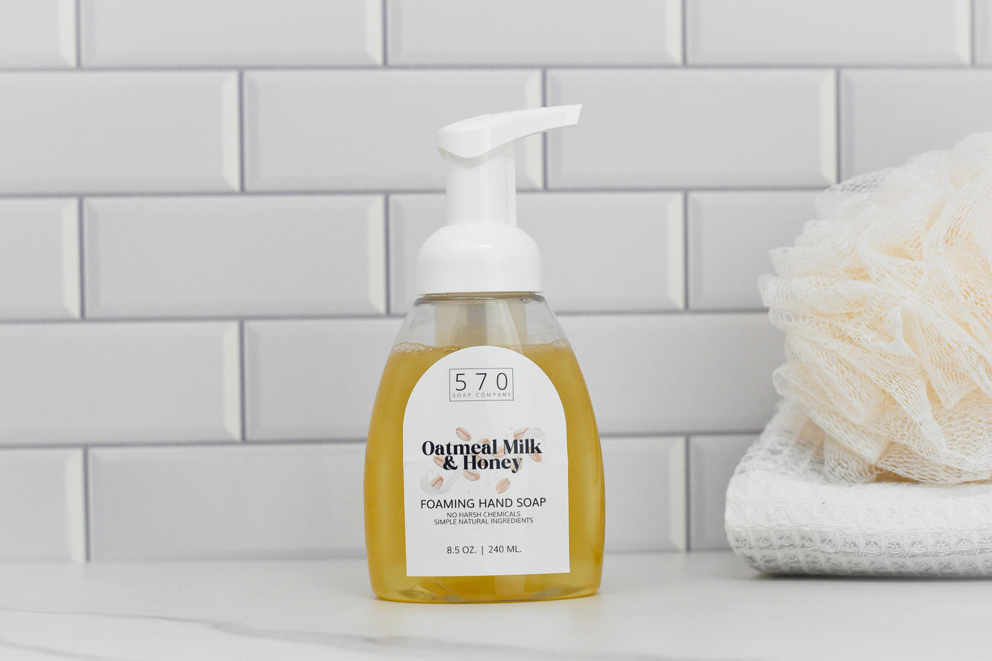 A bottle of 570 Soap Co.'s Oatmeal Milk & Honey Scented Foaming Hand Soap, complete with a white pump, sits on a bathroom counter. The background showcases white subway tiles, while a white loofah rests on a folded towel, emphasizing the soap's luxurious moisturizing benefits.