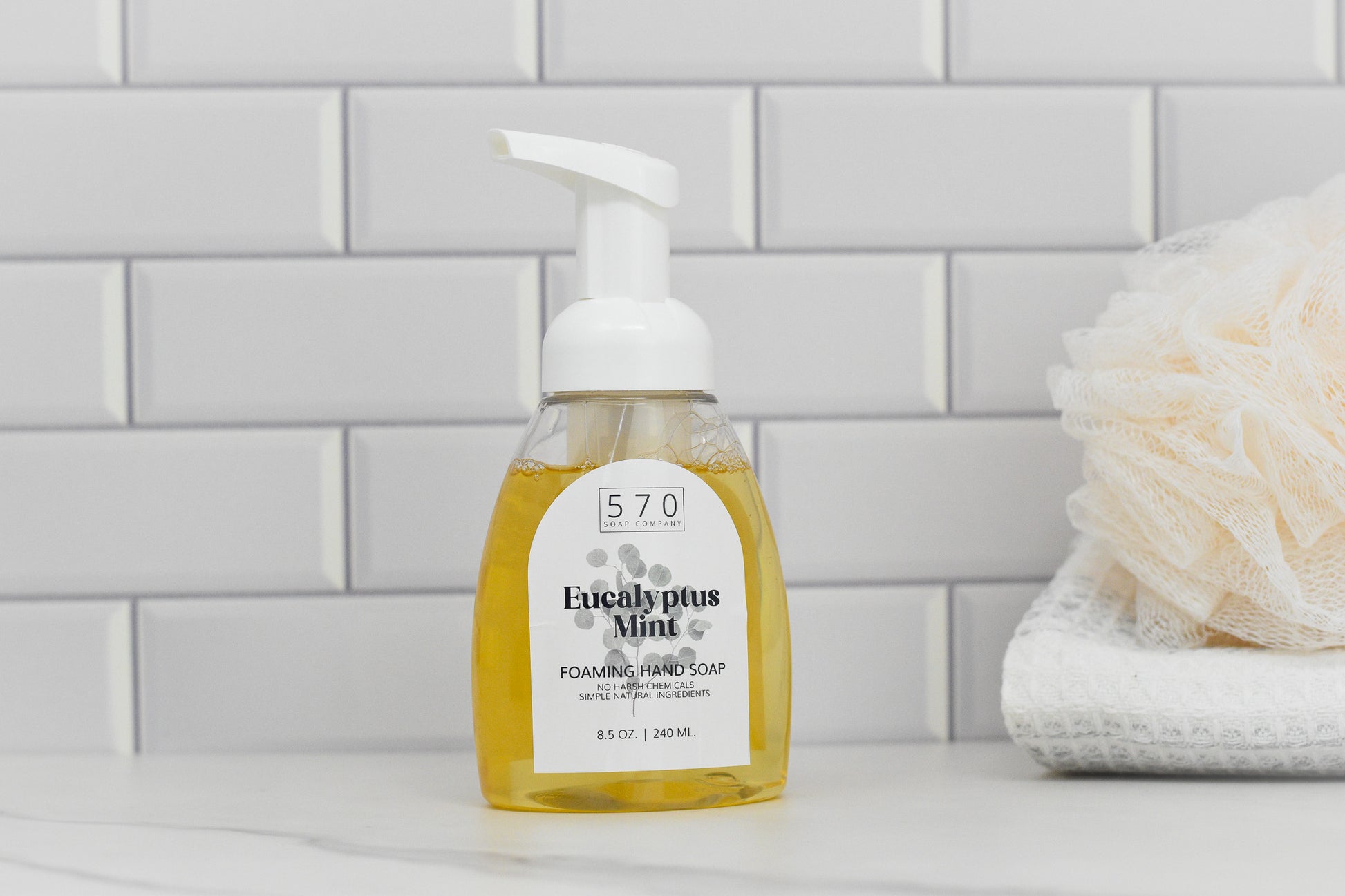 On the bathroom counter sits a bottle of 570 Soap Co.s Eucalyptus Mint All Natural Foaming Hand Soap, made with essential oils. In its clear pump bottle, its set against white tiled walls, with a beige bath sponge and folded towels partially visible on the right.