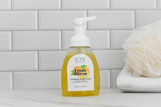 A bottle of 570 Soap Co.s Fresh Citrus All Natural Foaming Hand Soap sits on a bathroom countertop, infused with essential oils. In the background, a white loofah and folded towels are artfully arranged against a subway tile wall.