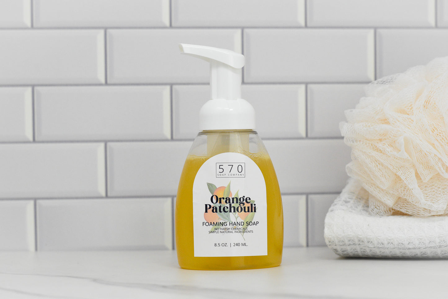 An Orange Patchouli Natural Foaming Hand Soap, made with essential oils by 570 Soap Co., sits on a bathroom counter against a white brick tile backsplash. Next to it, a folded towel and bath scrub await, promising an aromatic experience. The label indicates the volume of 8.5 oz (240 ml).