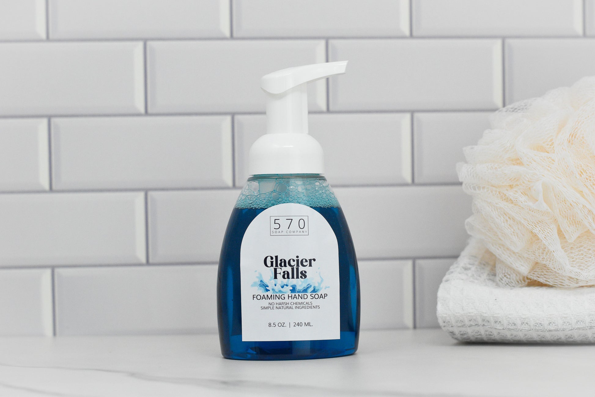 A bottle of 570 Soap Co.'s Glacier Falls Scented Foaming Hand Soap, known for its refreshing masculine aroma, rests on a white tiled surface, with its pump dispenser poised for use. Next to it, a neatly folded white towel is crowned with a bath sponge. In the background, sleek white subway tiles finish off the minimalist setting.