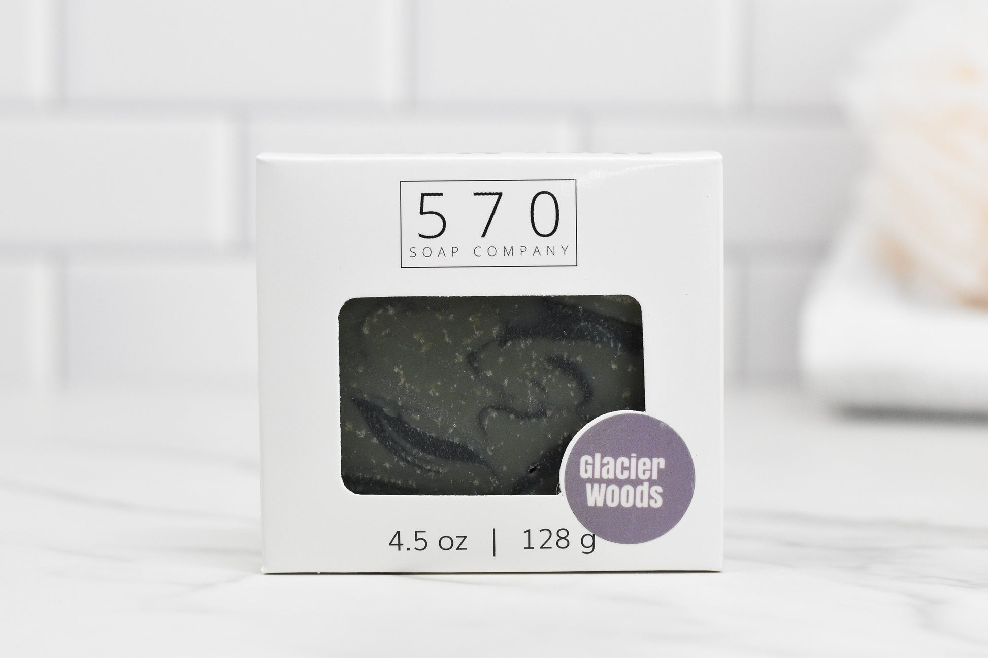 A box of Glacier Woods Bar Soap from 570 Soap Co. weighs 4.5 oz (128 g). The packaging, in white and featuring a window that showcases the dark scented soap inside, is elegantly displayed against a white tile background.