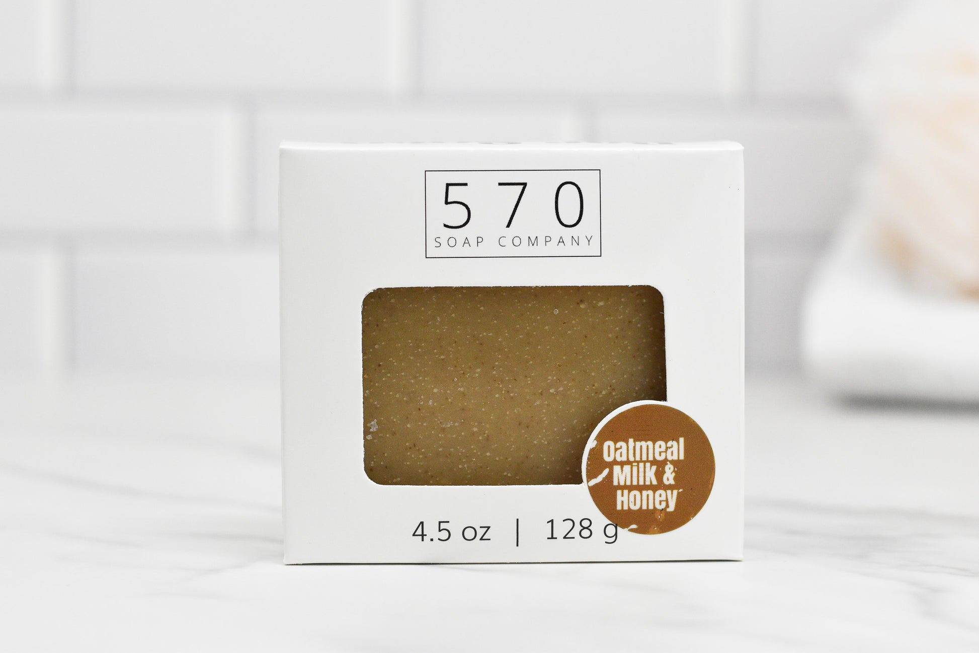 The Oatmeal Milk and Honey Bar Soap from 570 Soap Co. is showcased in a white box featuring a cutout that reveals the 4.5 oz (128 g) soap inside. This nourishing blend, set against a softly focused white tiled wall, beckons you to enjoy its rich creamy milk and sweet honey essence.