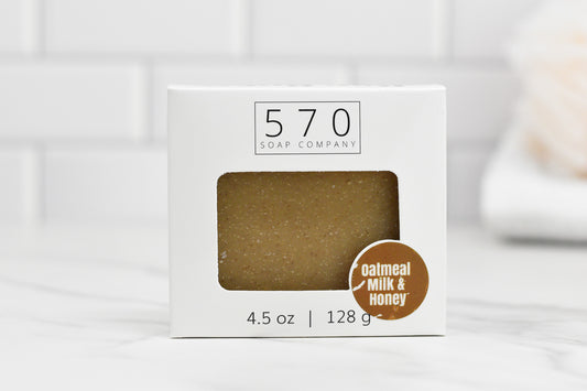 The Oatmeal Milk and Honey Bar Soap from 570 Soap Co. is showcased in a white box featuring a cutout that reveals the 4.5 oz (128 g) soap inside. This nourishing blend, set against a softly focused white tiled wall, beckons you to enjoy its rich creamy milk and sweet honey essence.