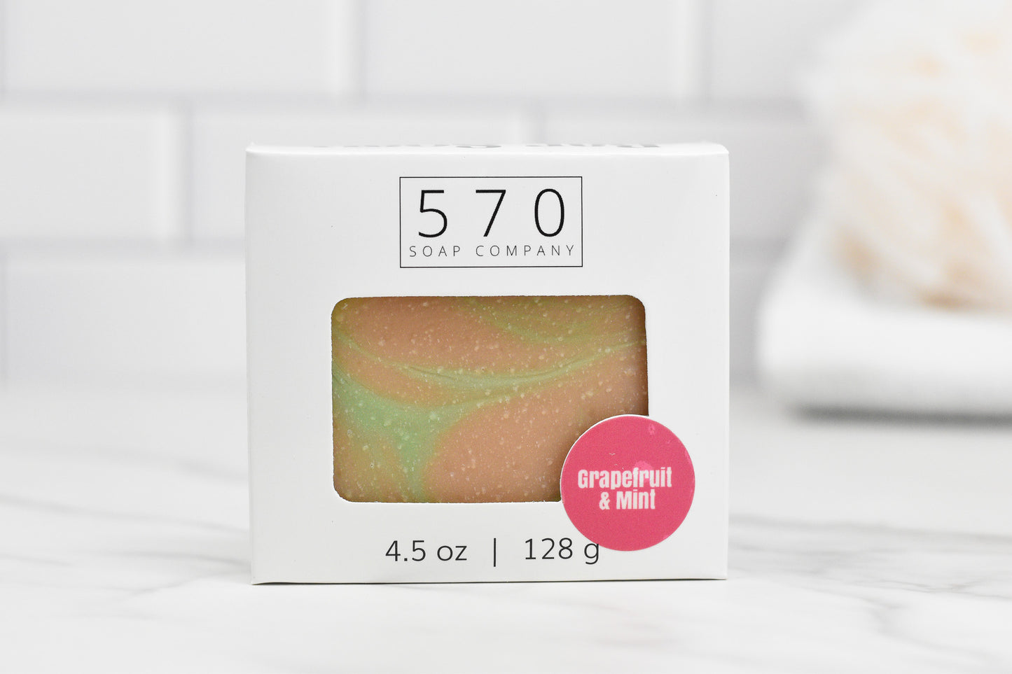 A boxed Fresh Grapefruit & Mint Bar Soap from 570 Soap Co. sits elegantly on a marble surface. The box features a transparent window revealing the multicolored soap inside. A pink sticker proudly showcases the enticing blend, promising a refreshing and invigorating experience.