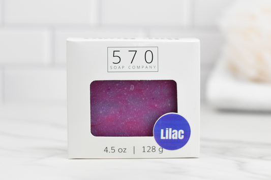 A box of 570 Soap Co.'s Luscious Lilac Bar Soap: Floral, enriched with a nourishing blend and floral scent, is showcased. The packaging includes a window showcasing the purple soap inside and indicates it weighs 4.5 oz (128 g). In the background, there is a softly focused view of a towel and white tiles.