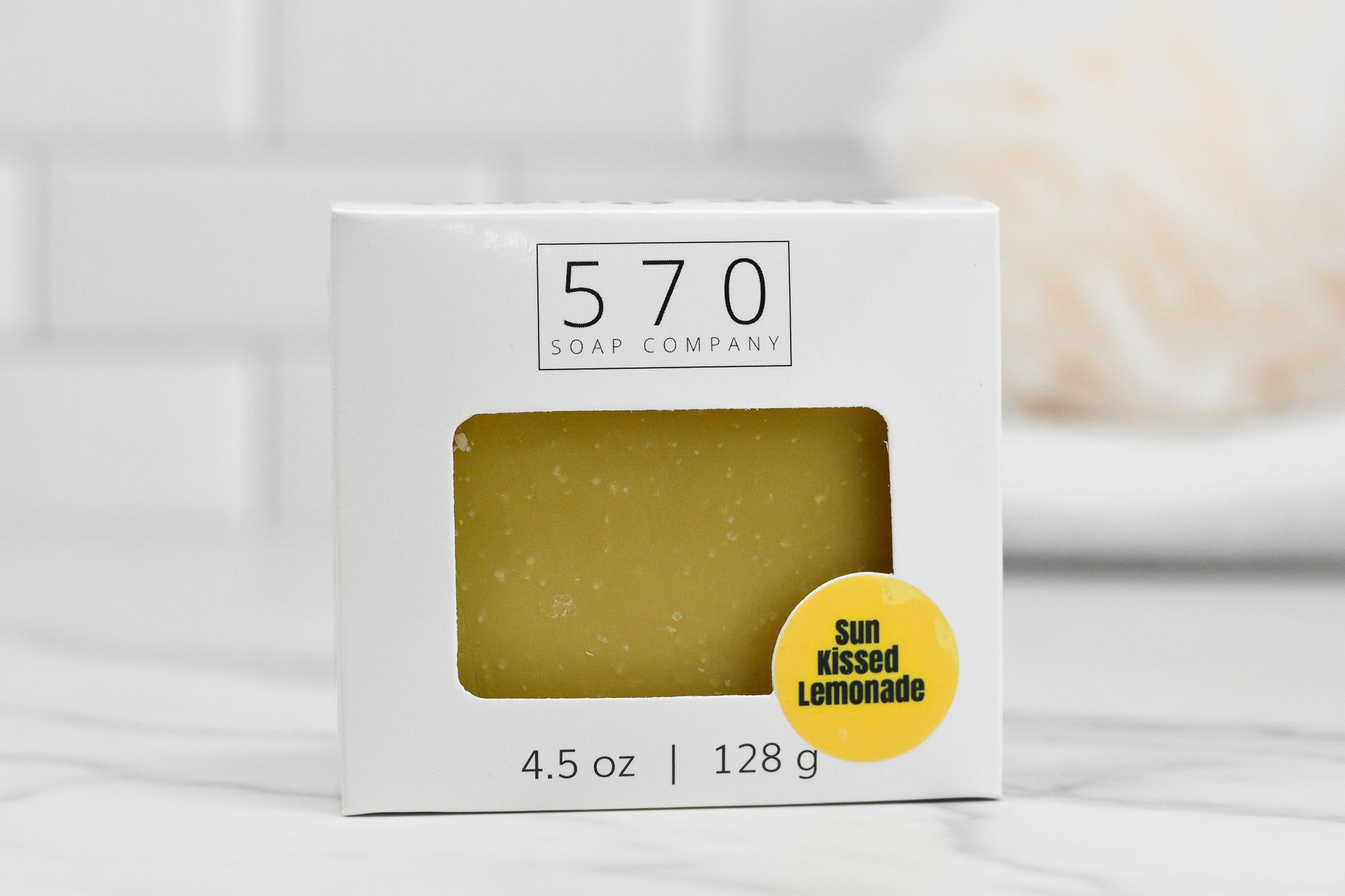 A 4.5 oz (128 g) box of Sun-Kissed Lemonade Bar Soap from 570 Soap Co. features a visible yellow bar inside. The label showcases "Sun Kissed Lemonade," emphasizing its citrus zest, and the package is set against a softly blurred white tiled wall, offering a promise of a refreshing cleanse.