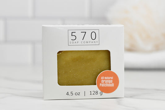 The Orange Patchouli Bar Soap - All Natural by 570 Soap Co. showcases a bar infused with orange patchouli essential oil. Housed in white packaging with a cut-out window, this 4.5 oz (128 g) soap adds an elegant touch to any bathroom setting in the background.