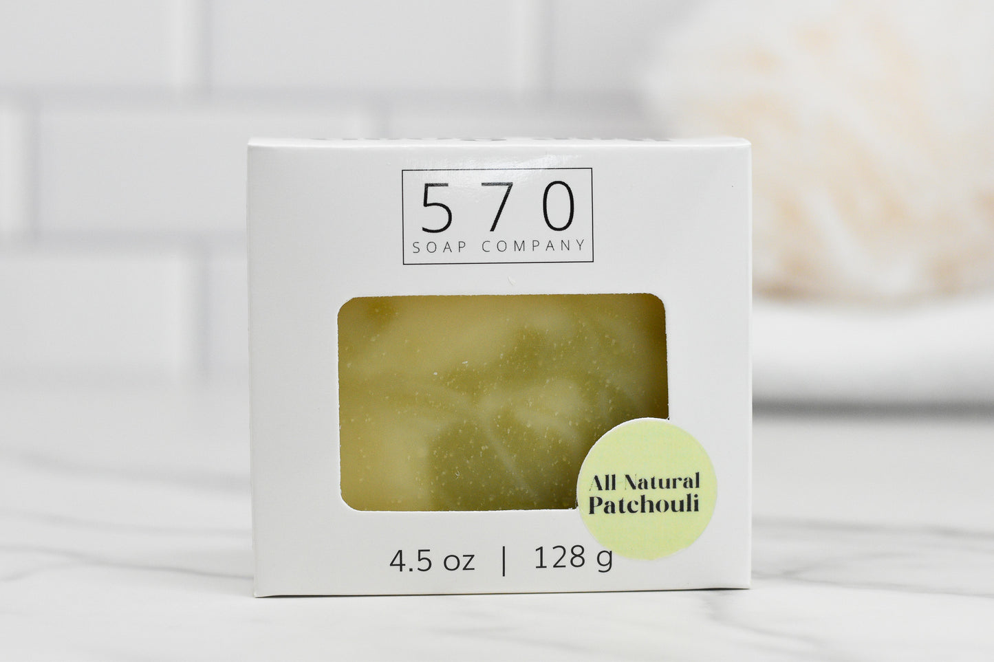 The Patchouli Essential Oil Bar Soap - All Natural from 570 Soap Co. exudes Earthy Elegance. The packaging, adorned in white and featuring a rectangular window, offers a peek at the soap inside. Labeled with "All Natural Patchouli" and enriched with Patchouli Essential Oil, it weighs 4.5 oz / 128 g against a tiled surface background.