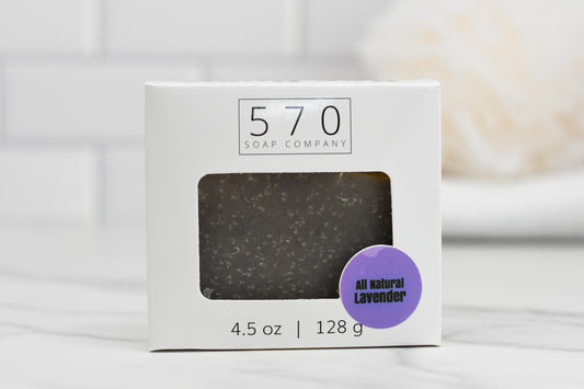 A bar of Lavender Essential Oil Bar Soap - All Natural from 570 Soap Co. comes in a white box featuring a clear window and adorned with a purple sticker that highlights the inclusion of lavender essential oil. The packaging indicates the soap weighs 4.5 oz | 128 g and rests on a light, glossy surface with a softly blurred background.