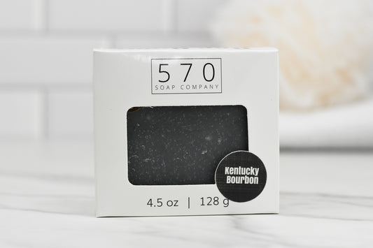 A bar of soap enclosed in a white box featuring a rectangular window highlights the Kentucky Bourbon Charcoal Bar Soap. Bearing the name 570 Soap Co., it radiates Southern elegance. Weighing 4.5 oz or 128 g, it sits gracefully on a white marble surface.