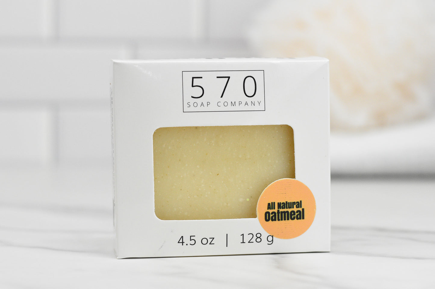 The Oatmeal Bar Soap - All Natural from 570 Soap Co. is enriched with coconut oil and comes packaged in a minimalist white box featuring a small clear window. Weighing 4.5 oz (128 g), it rests against softly blurred white tiles.