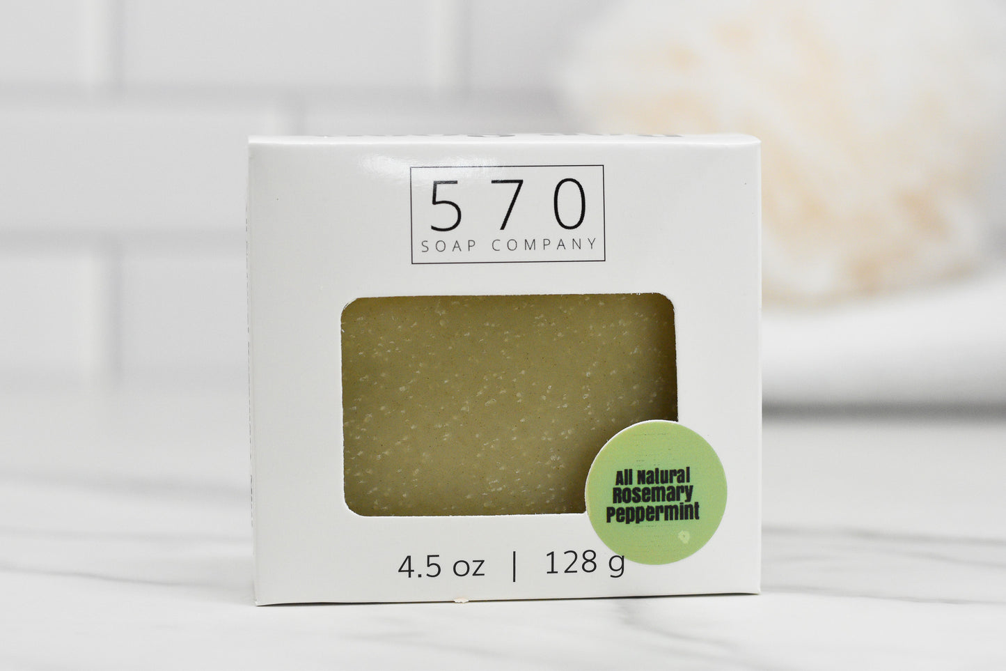 A boxed bar of Rosemary Peppermint Bar Soap labeled 570 Soap Co. with 4.5 oz | 128 g is featured. The packaging includes a green sticker that reads All Natural, emphasizing the use of rosemary peppermint essential oils in the soap. The background, a softly blurred bathroom setting, enhances its natural appeal.