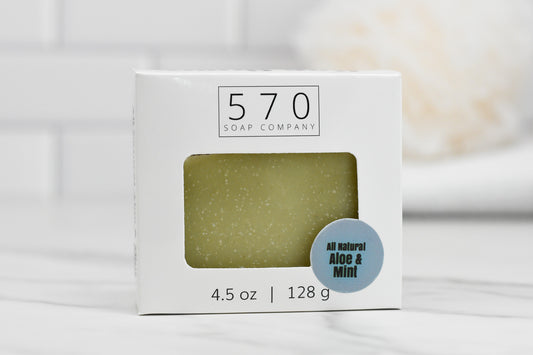 A white box with a cut-out window displays a green bar of Aloe & Mint Bar Soap - All Natural inside. The box is labeled 570 Soap Co. and features natural ingredients. It weighs 4.5 oz (128 g). The blurred background shows a white tiled wall.