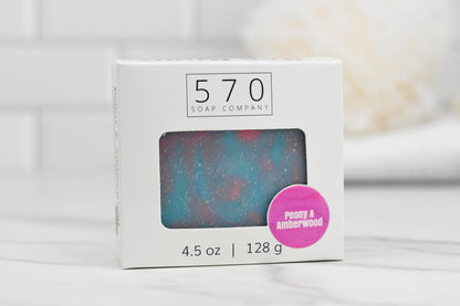 A rectangular box from 570 Soap Co. showcases a cut-out window revealing the Peony & Amberwood Bar Soap within. The front label emphasizes its enchanting fragrance and notes that the soap weighs 4.5 oz or 128 g. The backdrop of white tile enhances the product's appeal.