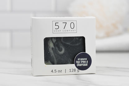 A box of Tea Tree & Charcoal Bar Soap - All Natural from 570 Soap Co. is displayed. The white packaging includes a clear window that reveals the soap inside, and a round label prominently showcases its all-natural ingredients: tea tree and activated charcoal. The soap weighs 4.5 oz (128 g).