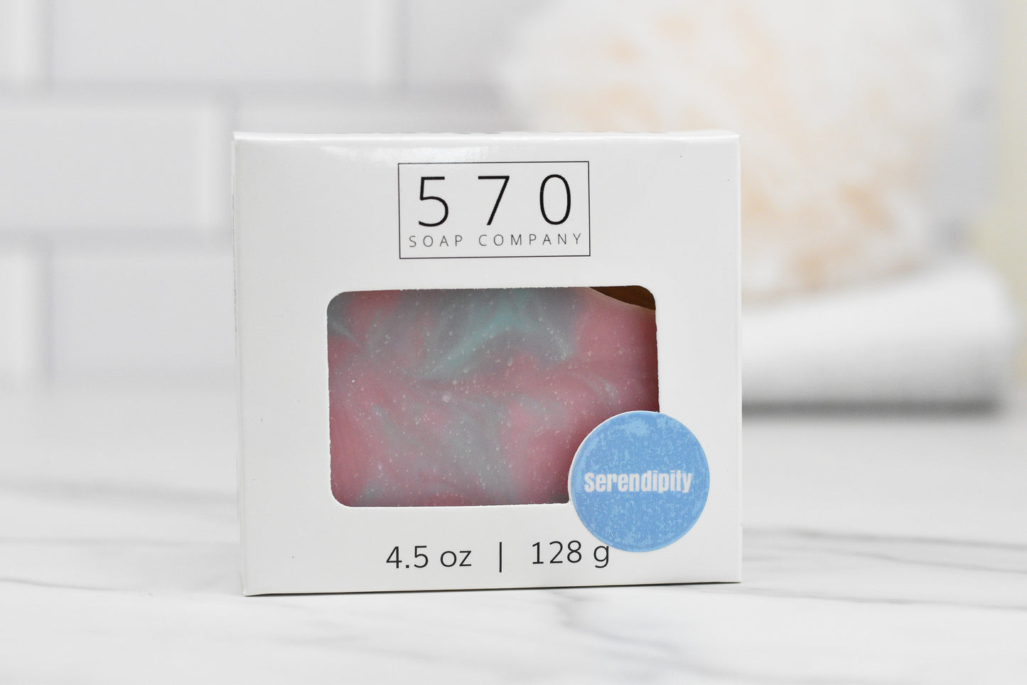 Displayed is a box of Serendipity Bar Soap from 570 Soap Co. This pink and blue marbled soap, weighing 4.5 oz or 128 g, features a moisturizing formula. Presented in a clean, light kitchen or bathroom setting, it ensures both elegance and hydration for your daily routine.