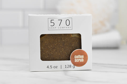 A box of 570 Soap Co. Coffee Scrub Bar Soap: Wood Shelf Cafe Coffee, enriched with exfoliating kaolin clay, weighing 4.5 oz (128 g), reveals a visible section of the brown, speckled soap within. The packaging is placed on a light surface against a blurred background.