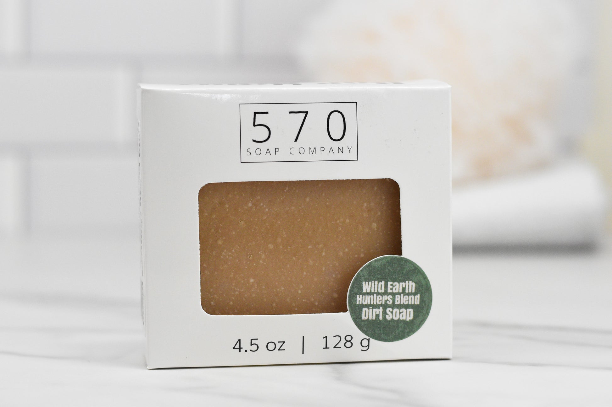 The Wild Earth Hunters Blend Bar Soap - Dirt Soap from 570 Soap Co. exudes a distinctive dirt-scented fragrance and is enriched with kaolin clay. Weighing in at 4.5 oz (128 g), it has a naturally earthy look, beautifully encased in a white box and showcased on a white marble surface.