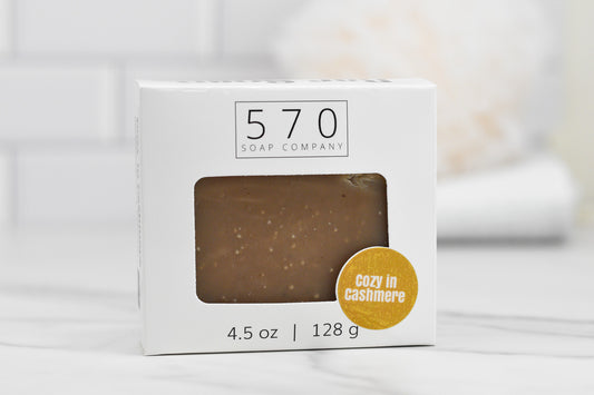 A bar of Cozy in Cashmere Bar Soap: Luxurious Blend from 570 Soap Co. sits in a white box featuring the words "Cozy in Cashmere" within a yellow circle. Placed on a white marble surface, its aromatic blend is sure to enchant the senses against the gently blurred background.