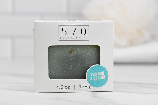 A white box of Sea Salt & Orchid Bar Soap: Coastal Tranquility, branded by 570 Soap Co., features a clear window that reveals the luxurious lather bar soap within. A blue sticker highlights "Sea Salt & Orchid." The background is a softly blurred bathroom setting with an immaculate white towel.