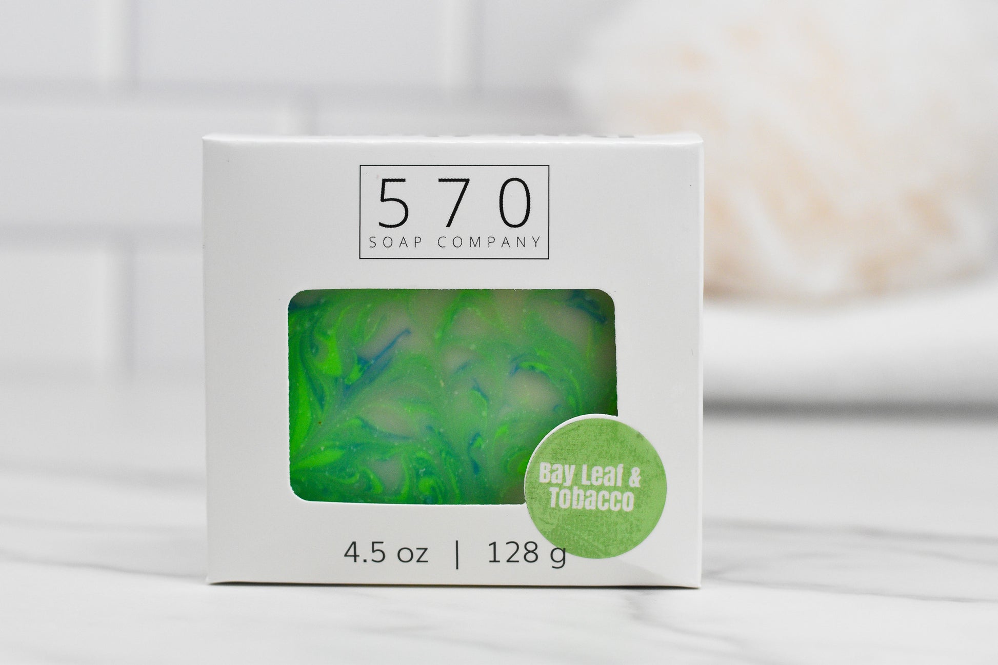 A soap box from 570 Soap Co. displays the Bay Leaf and Tobacco Bar Soap, featuring a green and blue marbled design. The base of the box indicates 4.5 oz | 128 g, while a circular label emphasizes its bay leaf and tobacco scents. A blurred bathroom setting in the background enhances its enticing presentation.