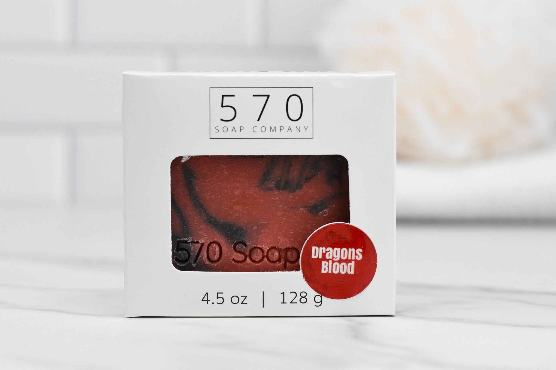 A box of Dragons Blood Bar Soap from 570 Soap Co. showcases an exotic fragrance. The packaging is white with a clear window that reveals the red handcrafted soap within. Weighing 4.5 oz (128 g), it is positioned on a white, tiled surface with a blurred towel in the background.