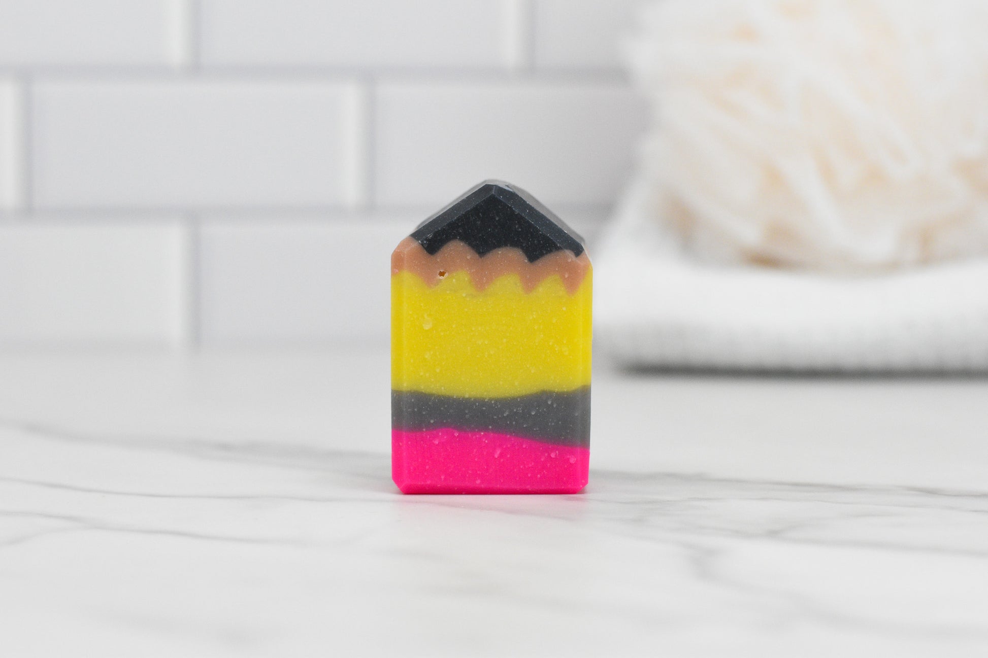 The Cute Pencil Shaped Bar Soap by 570 Soap Co., ideal for gifting to teachers, boasts vibrant layers of black, yellow, brown, and pink against a white marble backdrop. The soft-focused background hints at a bathroom environment with white tiles and a neatly folded towel.