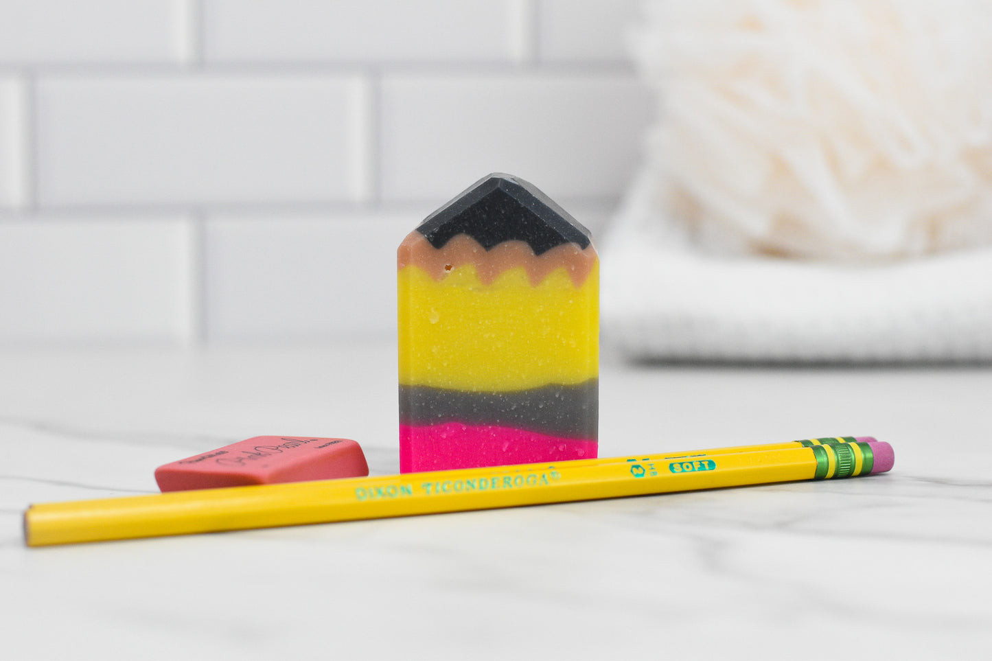 A multicolored bar of the Cute Pencil Shaped Bar Soap from 570 Soap Co. rests on a marble surface, next to a sharpened yellow pencil and an orange eraser. The white, textured background with blurred elements suggests a bathroom or classroom setting, making it an ideal back-to-school surprise or teachers’ gift.