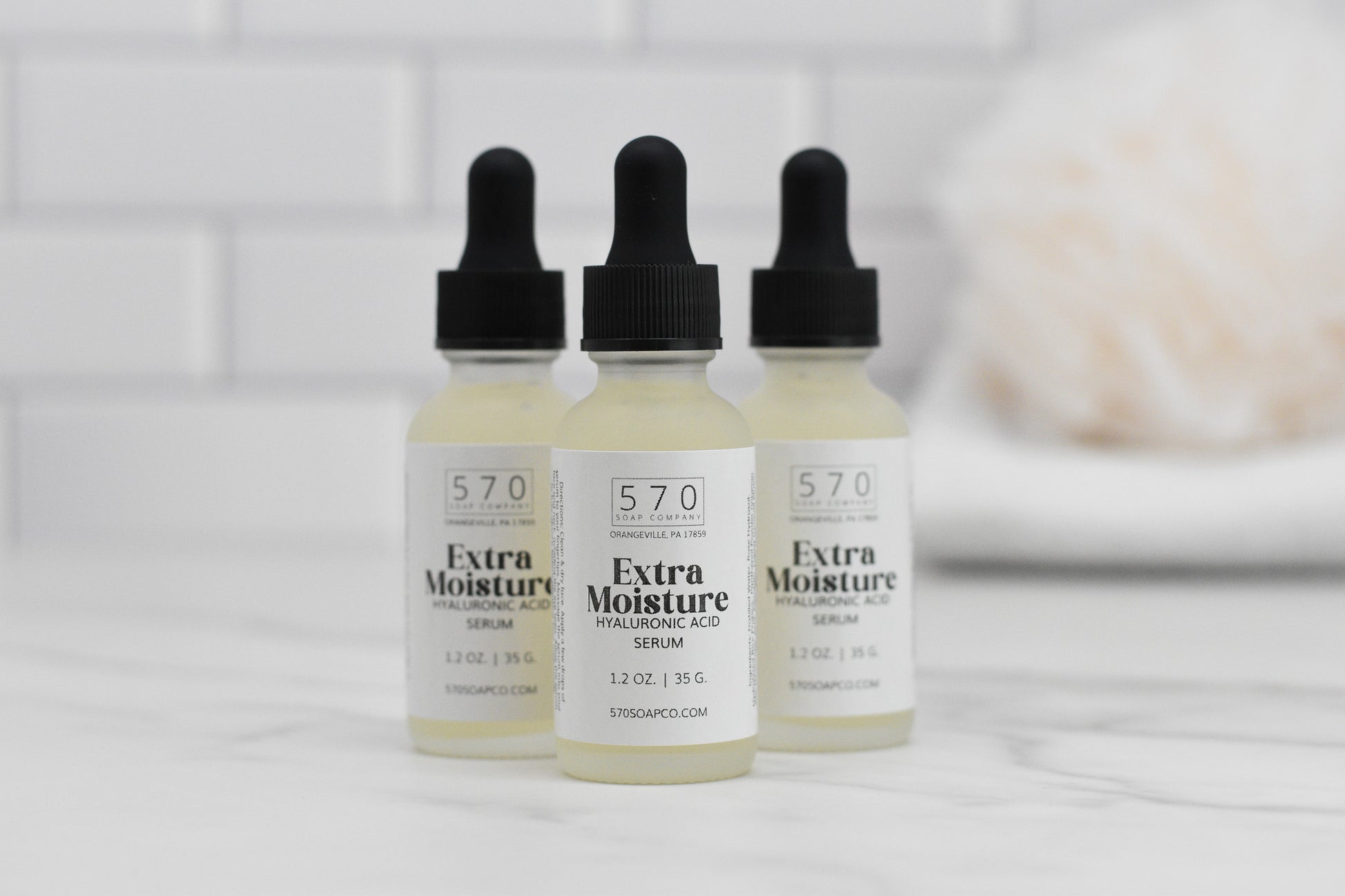 Three bottles of 570 Soap Co.'s Extra Moisturizing Hyaluronic Acid Serum are elegantly displayed on a white marble surface. With sleek black dropper caps, they promise to reduce fine lines by delivering intense hydration. The background is adorned with white subway tiles, accompanied by a softly focused object that resembles a towel.