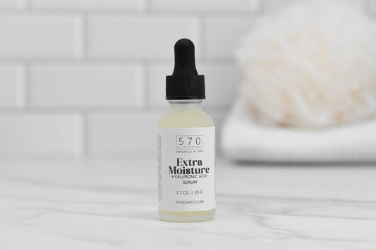 A small bottle with a dropper labeled Extra Moisturizing Hyaluronic Acid Serum by 570 Soap Co. sits on a marble surface. Offering intense hydration, it's ideal for reducing fine lines. In the background, blurred white tiles and a folded towel can be seen.