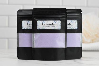 Three black pouches of 570 Soap Co.'s Lavender Fizzing Bath Dust sit on a white surface against a tiled background. Each pouch features a clear window revealing the light purple powder inside, offering an invitation to relax and enjoy a customizable bath experience.