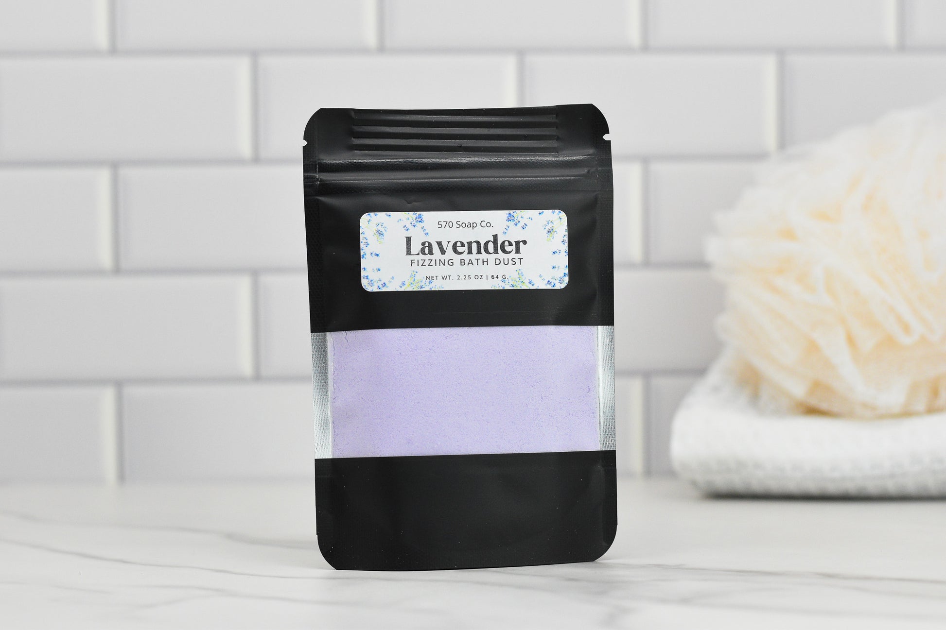 A black pouch of 570 Soap Co.'s Lavender Fizzing Bath Dust is elegantly placed on a bathroom counter against a white tile backdrop. Beside it rests a cream-colored bath sponge atop a white towel, promising customizable use for ultimate relaxation.