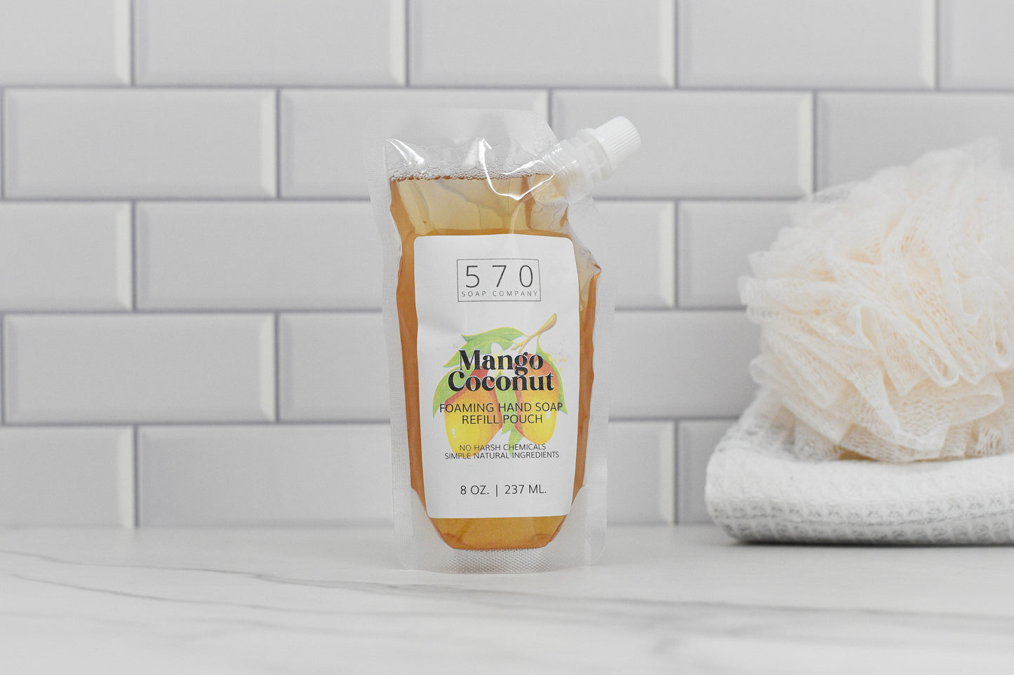 A pouch of Mango Coconut Scented Foaming Hand Soap from 570 Soap Co. sits on a white marble surface, beside folded towels and a bath sponge. Made with natural ingredients, this soap enhances the clean look of white subway tiles.