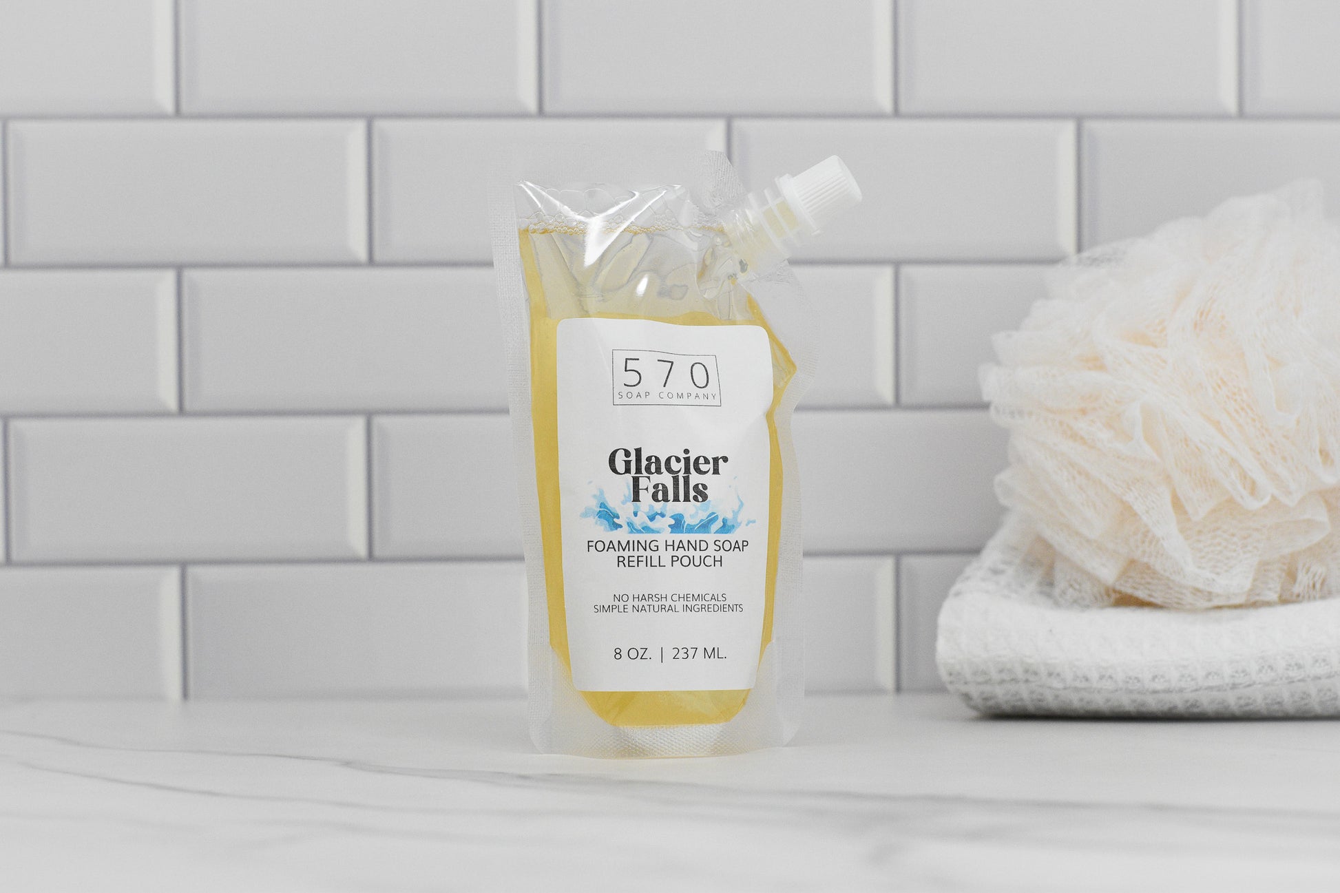 A refill pouch of 570 Soap Co.'s Glacier Falls Scented Foaming Hand Soap, known for its masculine fragrance and natural ingredients, rests on a bathroom counter. The clear pouch with a white label is accompanied by a white shower puff and towels in the background against a tiled wall.