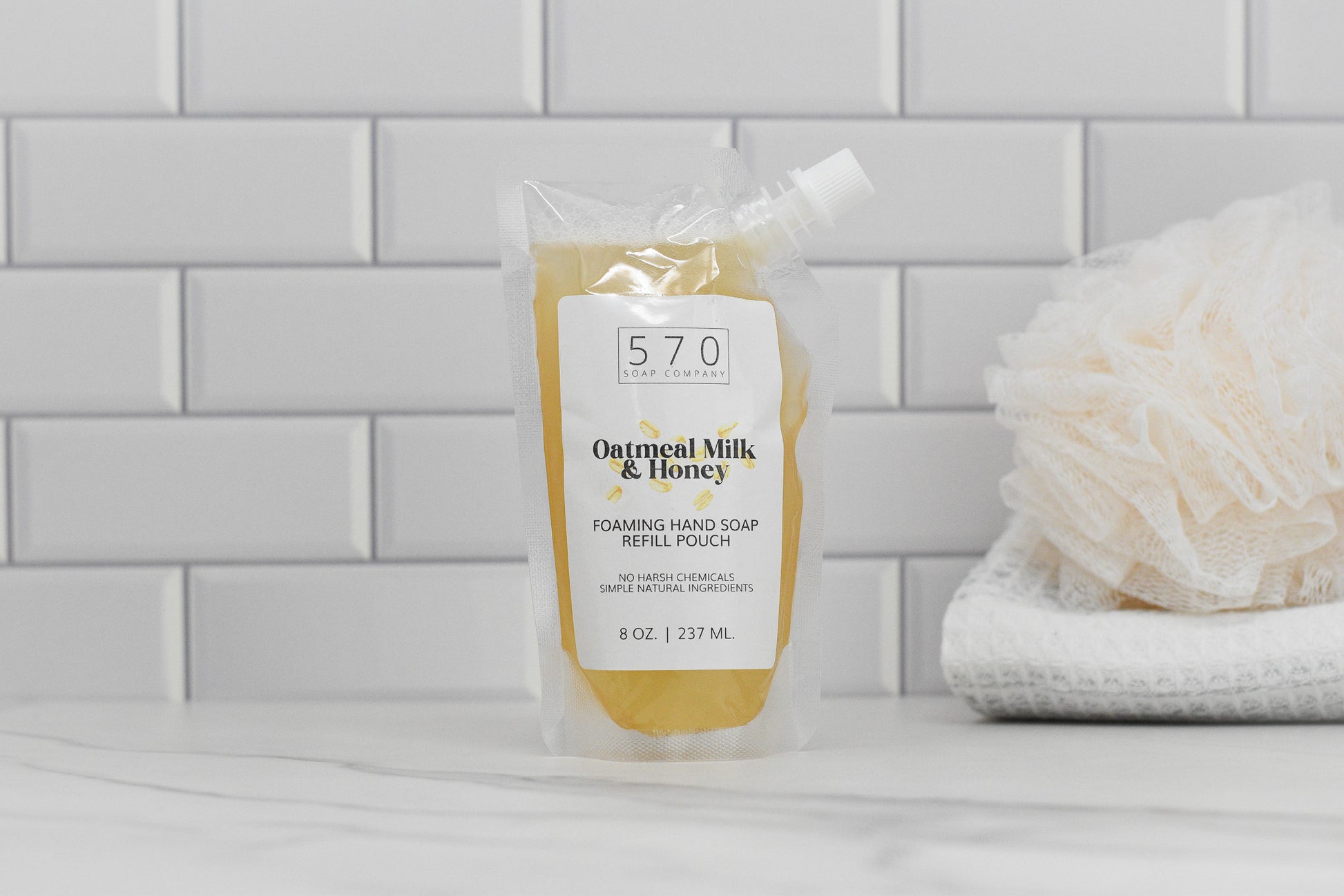 A refill pouch of Oatmeal Milk & Honey Scented Foaming Hand Soap from 570 Soap Co. is displayed on a white countertop, providing moisturizing benefits. In the background, a white loofah and folded towel rest against a tiled wall.